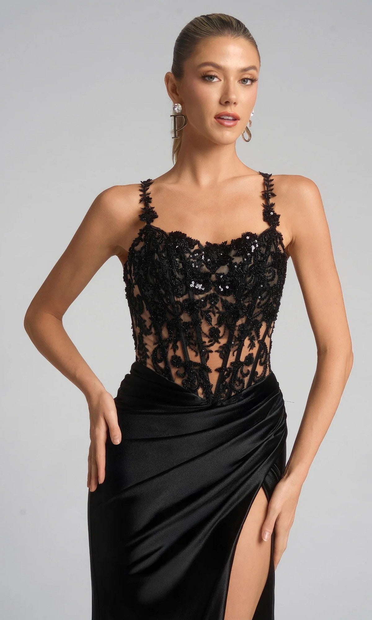 A woman wearing the Portia and Scarlett XO119 long formal dress, which has a black lace embroidered corset bodice, spaghetti straps, and a high slit in its flowing satin skirt.